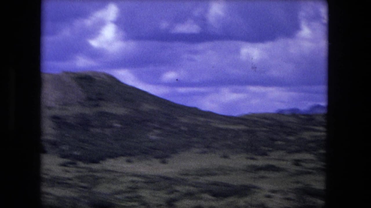 Mountain slope and rolling terrain beneath heavy clouds, framed by black film borders.
