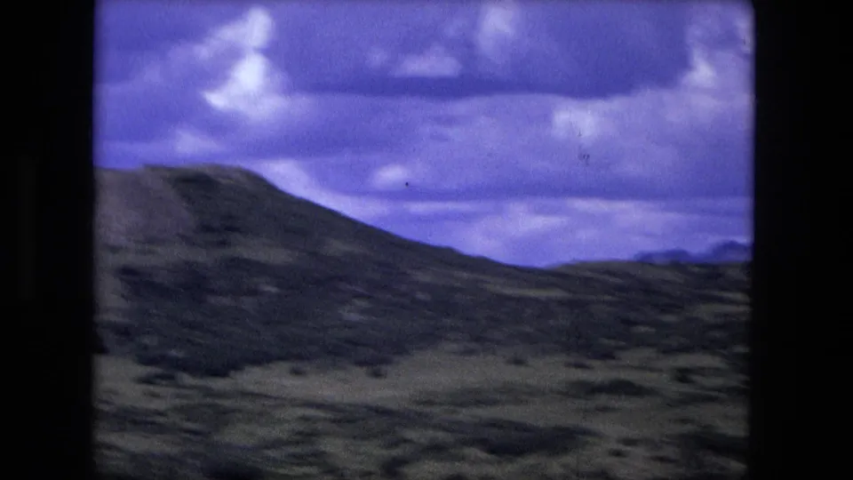 1976 8mm travel view of a broad mountain slope and rolling scrubland beneath dramatic cloud cover, likely filmed in motion.