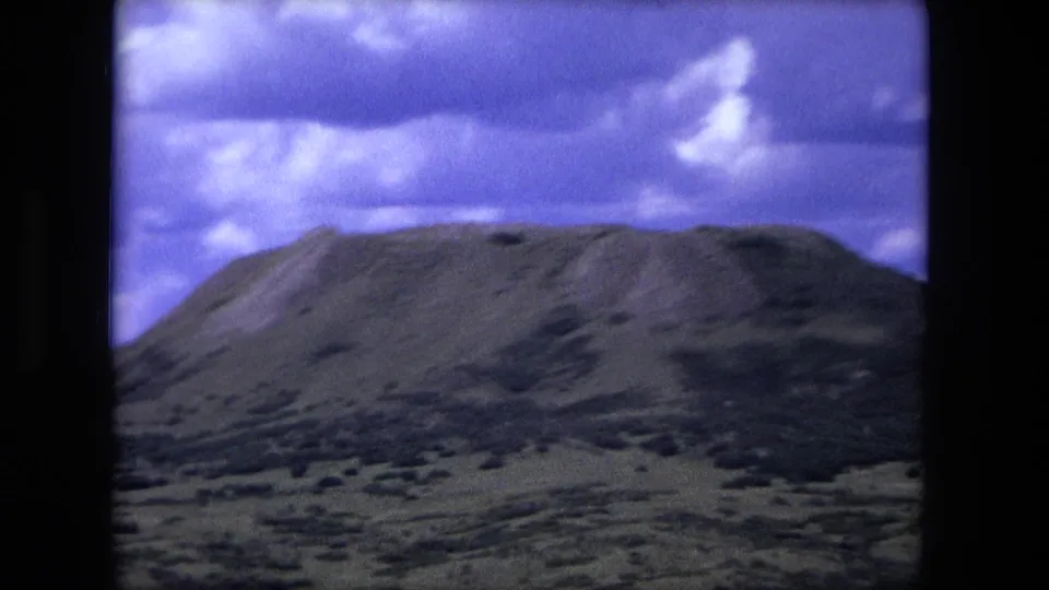 1976 8mm frame showing a rounded hill and scrub-covered slopes under bright cloud breaks and a purple-blue sky.