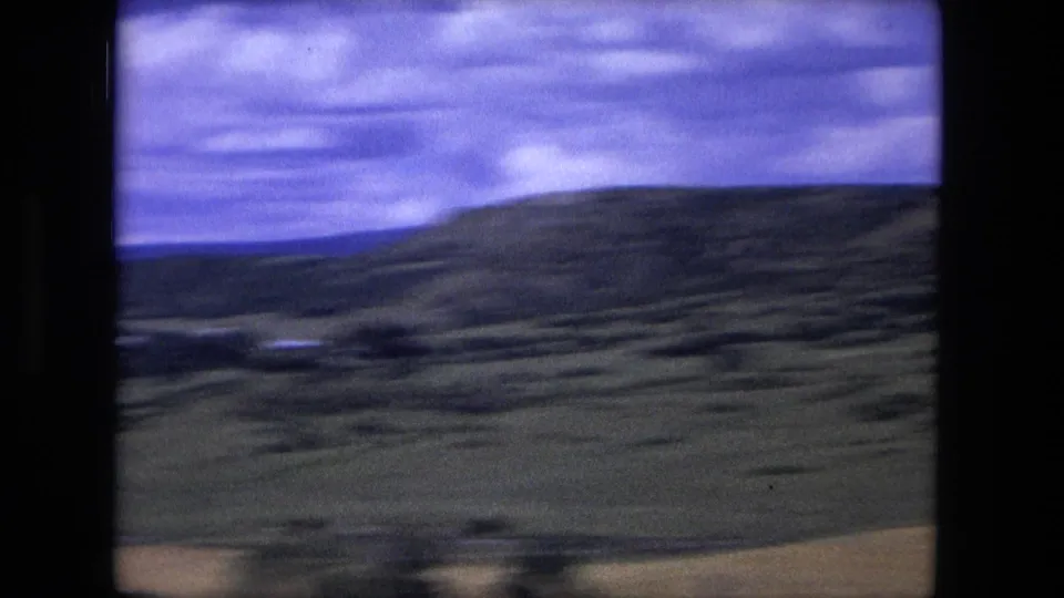1976 8mm travel frame showing rolling hills, open land, layered clouds, and a small bright feature in the valley distance.