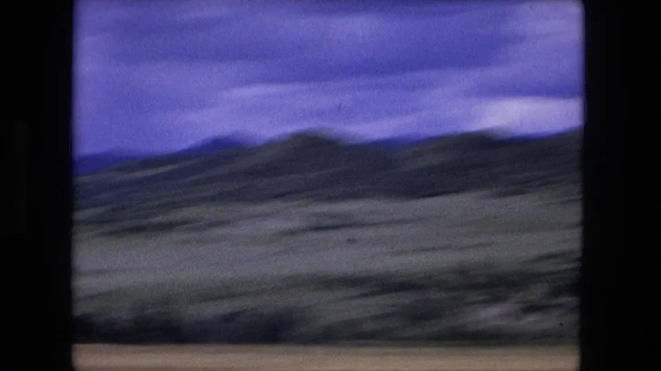 A moving 8mm view across sloped rural terrain and low hills, with blur suggesting the camera is inside a traveling vehicle.