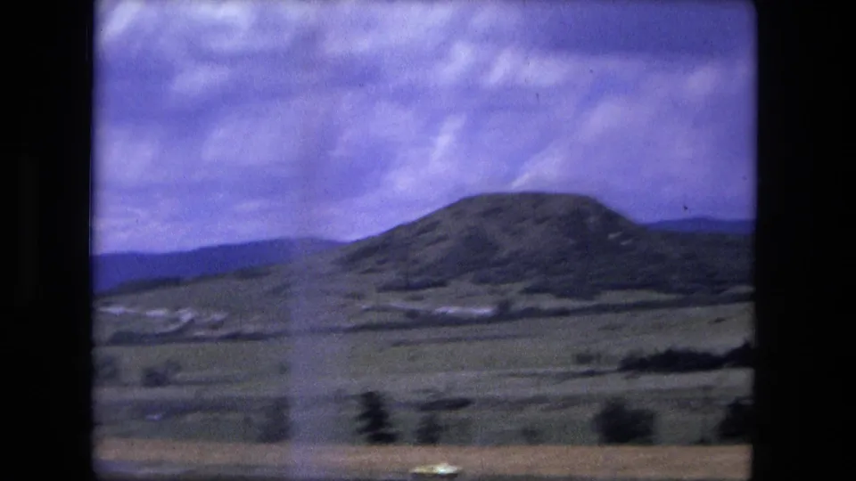 1976 home-movie frame showing a rounded hill, patchy fields, and distant ridgelines filmed from the open road.