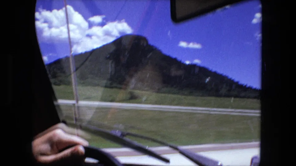 Interior road view with a visible hand on the steering wheel, windshield wipers, road, and a prominent hill ahead.