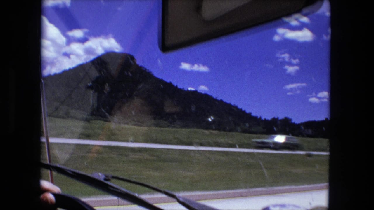 Passenger-seat view through a windshield toward a highway, grassy slope, hills, and blue sky.