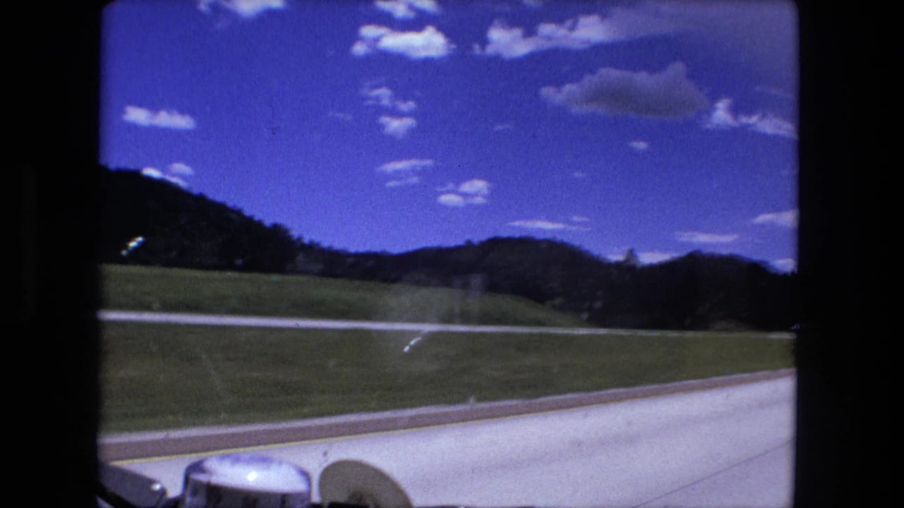 A Fort Wayne-area 1976 car-window view shows a paved roadside, grassy fields, low hills, clouds, and part of the dashboard.