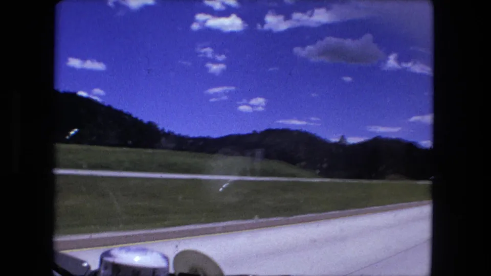 A Fort Wayne-area 1976 car-window view shows a paved roadside, grassy fields, low hills, clouds, and part of the dashboard.
