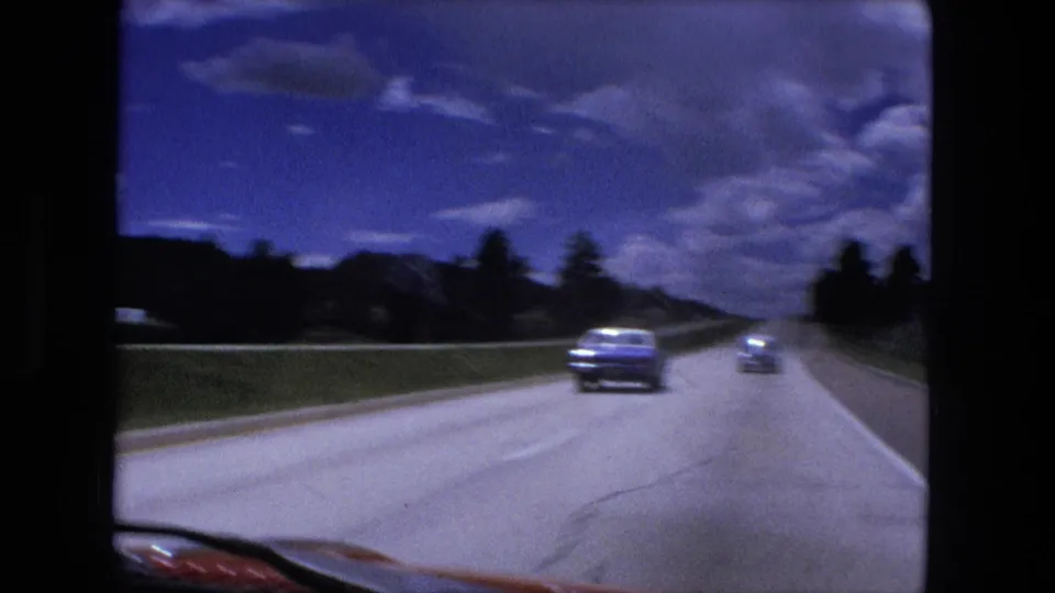 1976 8mm highway view near Fort Wayne, with cars ahead on the road, roadside trees, distant hills, and visible windshield edges.