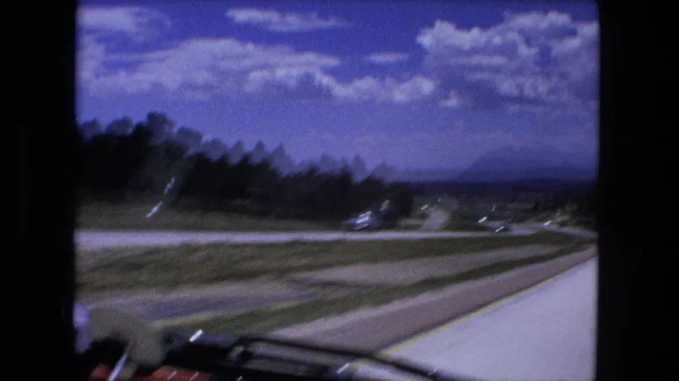 The passenger-side camera looks across a curving highway, grassy median, distant hills, sky, and car interior edges in this 1976 home movie frame.