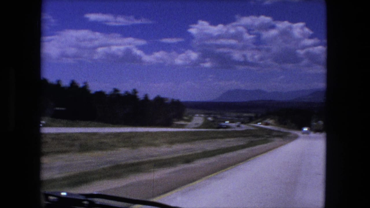 1976 8mm windshield view in Fort Wayne, Indiana, showing a curving highway, open fields, trees, and distant traffic.