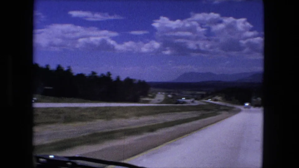 1976 8mm windshield view in Fort Wayne, Indiana, showing a curving highway, open fields, trees, and distant traffic.