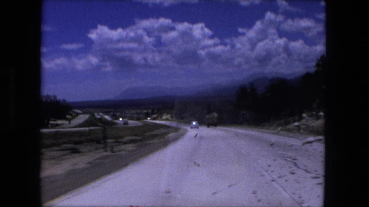 Fort Wayne, Indiana road-travel frame from 1976 with a pale highway, tree line, open roadside, and small vehicles ahead.