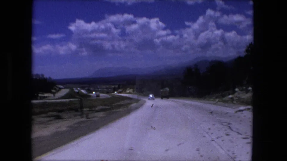 Fort Wayne, Indiana road-travel frame from 1976 with a pale highway, tree line, open roadside, and small vehicles ahead.