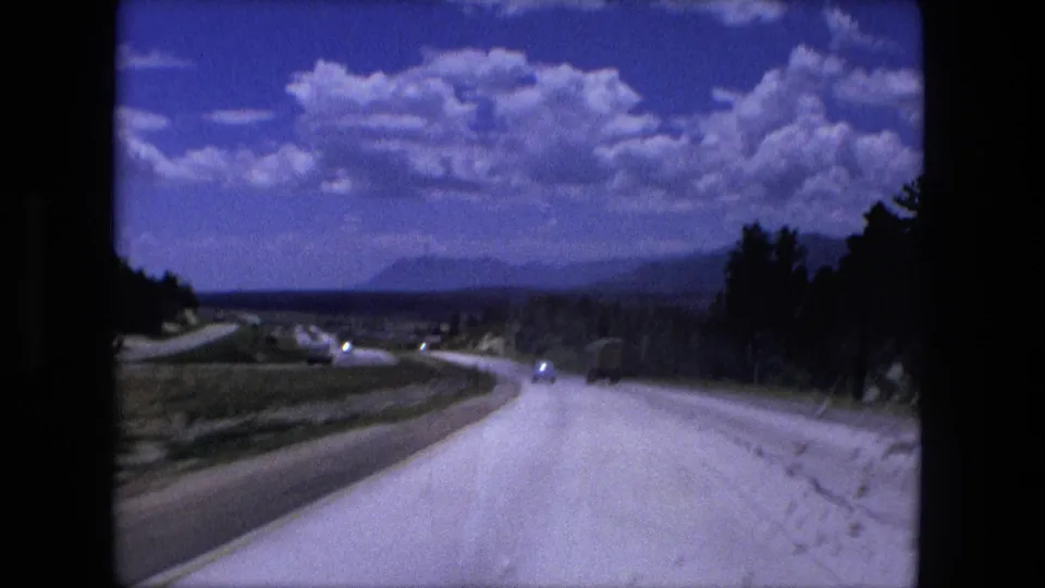 1976 8mm travel shot showing a forward view along a bright highway near Fort Wayne with trees, fields, and distant cars.