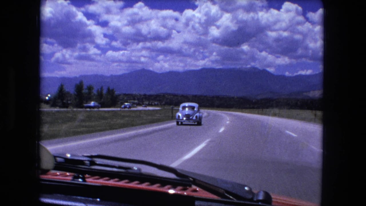 1976 8mm home-movie view from inside a red car traveling on a four-lane highway with a pale sedan ahead and distant hills under large clouds.