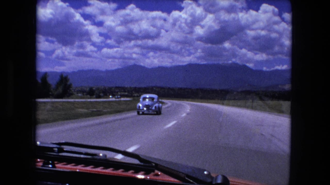 Highway curves gently ahead with a pale sedan in front, roadside fields, distant ridgeline, and bright clouds.