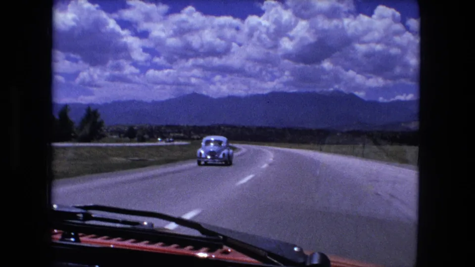 1976 in-car highway view showing a pale sedan ahead on a curving four-lane road, with fields and a distant ridgeline below a clouded sky.