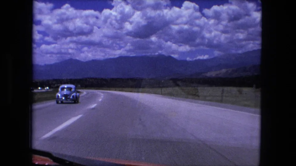 1976 8mm windshield view from a red car following a pale sedan on a wide rural highway, with open land and distant hills under heavy clouds.