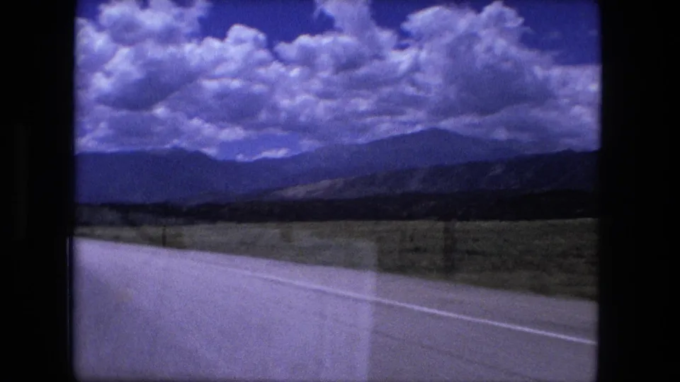 1976 8mm road-travel frame looking past the highway edge toward open fields, distant hills, and a broad sky of bright clouds.