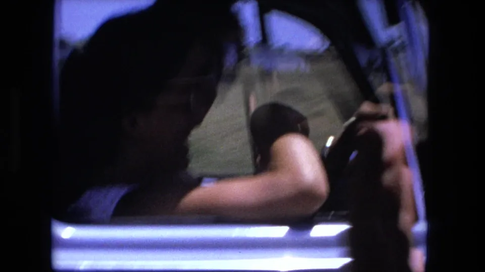 Inside the moving car, an adult driver is silhouetted through the open side window while holding the steering wheel during the 1970s road sequence.