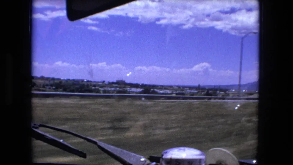 A passenger-side windshield view shows roadside grass, a distant highway area, low hills, and bright clouds during the Fort Wayne road-trip footage.