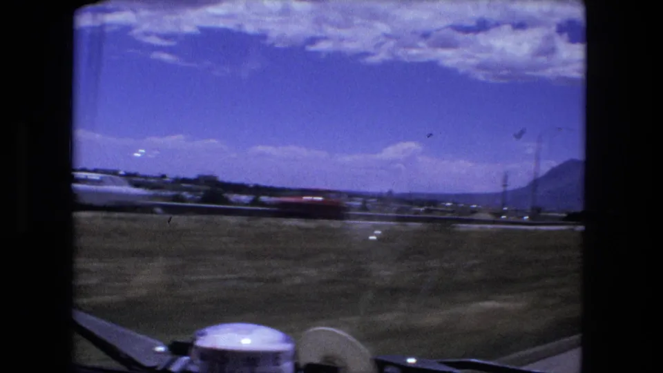 The final frame looks through the car windshield across open roadside ground, distant roadway structures, utility poles, and low hills under a bright sky.