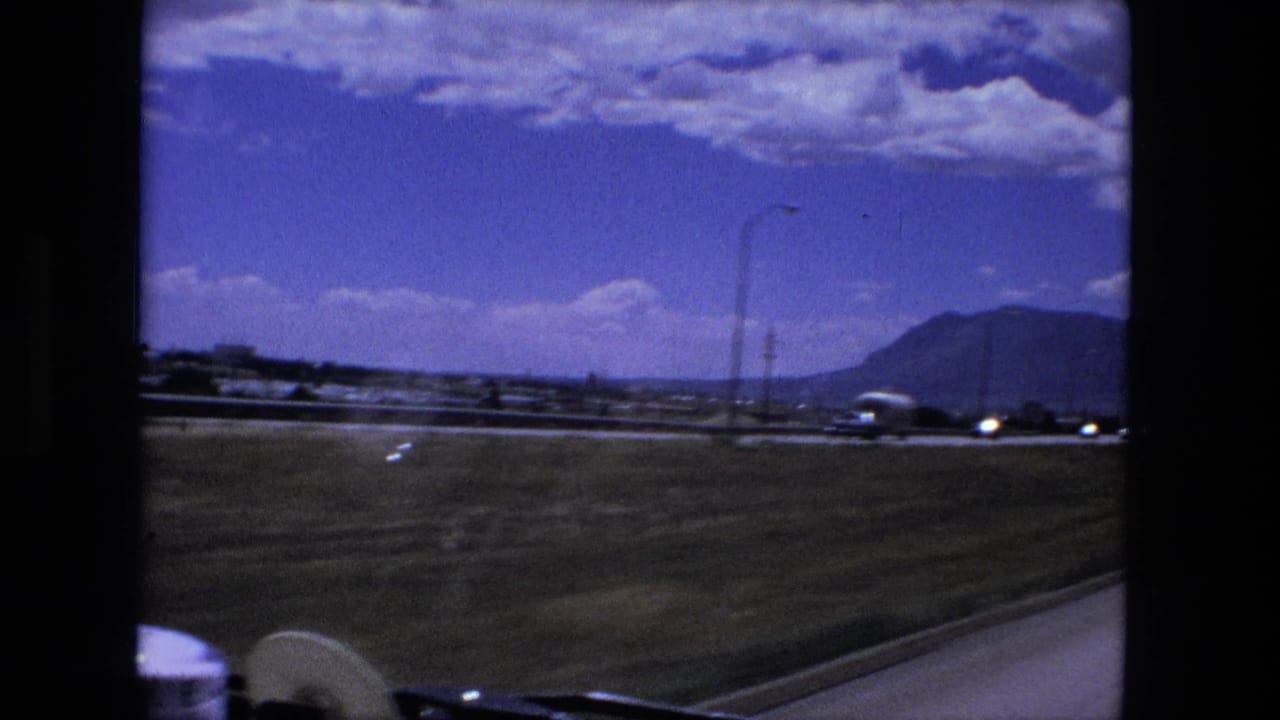 1976 8mm road-travel frame showing a highway scene near Fort Wayne metadata, with cars, open grass, distant hills, and a bright clouded sky.