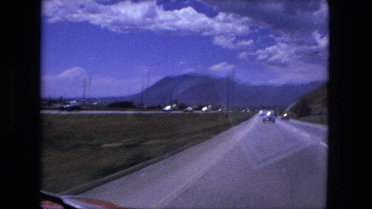 Forward view down a two-lane road with cars ahead, grassy roadside, hills, and a clouded sky.