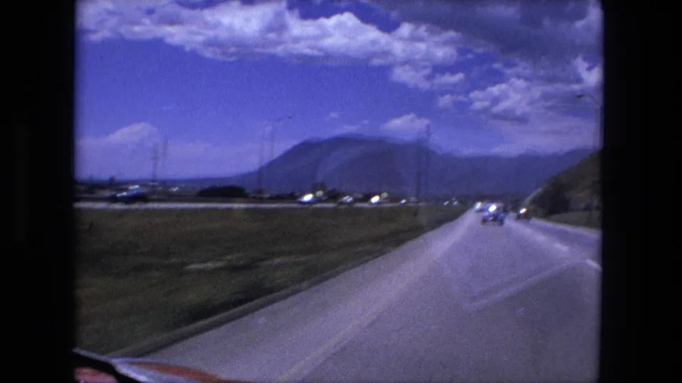 Forward-facing 1976 8mm driving view with cars on the road, a grassy shoulder, distant hills, and scattered clouds in the sky.