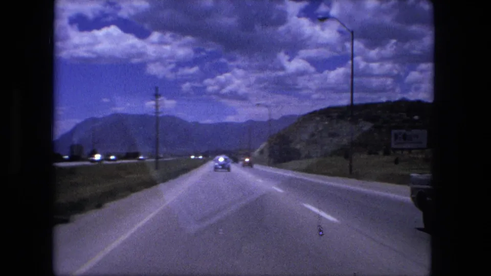 1976 8mm road scene from a moving car, showing highway lanes, traffic, utility poles, a roadside sign, distant hills, and heavy clouds.
