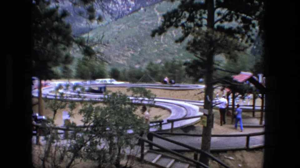 1976 8mm frame showing a roadside stop or overlook area with trees, fencing, curving pavement, parked cars, and small visible visitors.