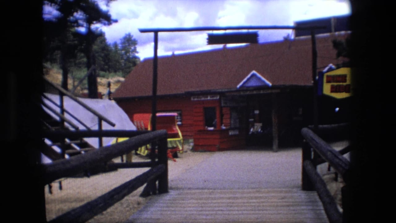 1976 Fort Wayne home-movie frame showing wooden railings approaching a red amusement park entrance building under a cloudy sky.
