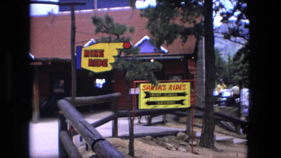 1976 Fort Wayne frame focusing on yellow attraction signs, wooden entrance rails, pine trees, and a rustic amusement park building.