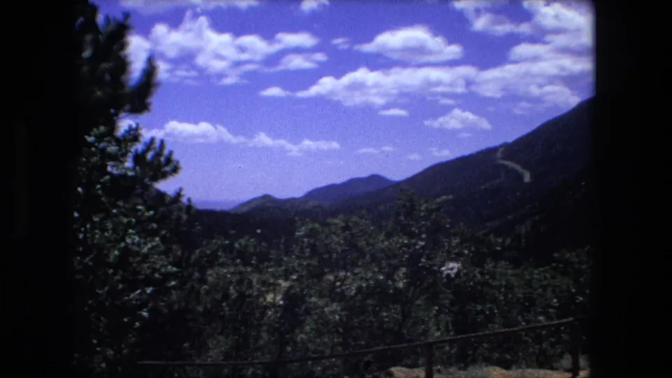 Archival 8mm view of wooded hills and a distant mountain ridge beneath a bright clouded sky.