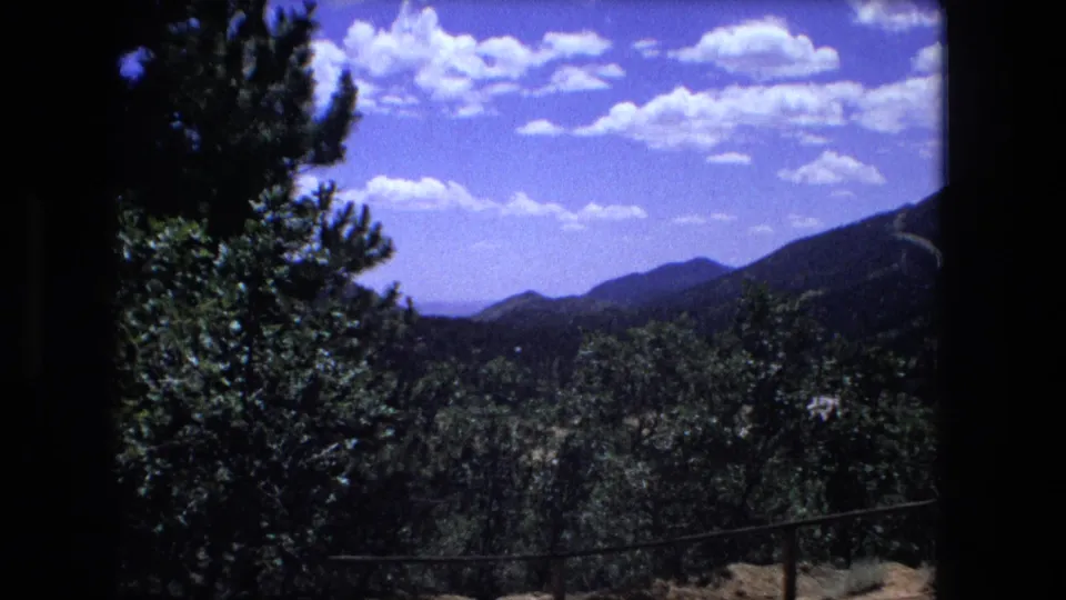 1970s home-movie frame showing a wooded mountain valley and distant ridges under scattered clouds.