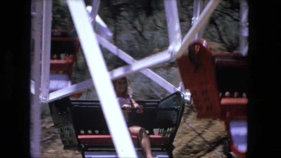 1976 8mm frame of a girl riding a small Ferris wheel seat, partly obscured by white metal supports.