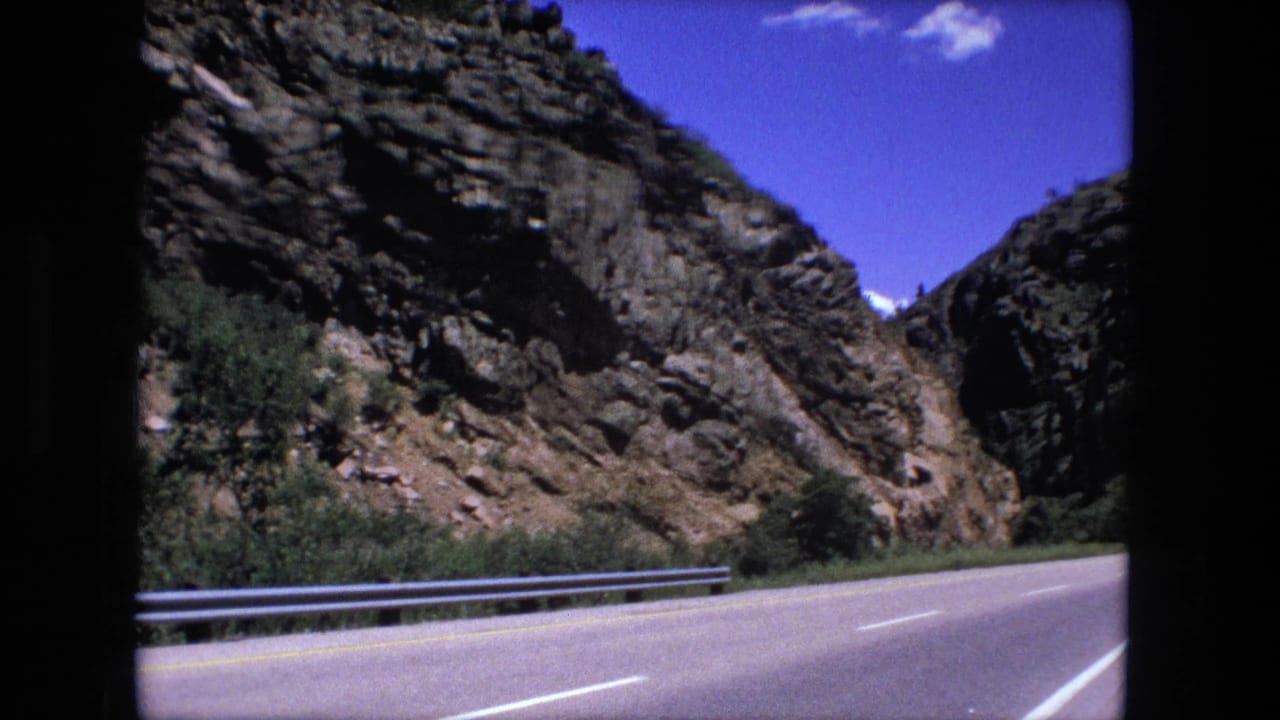 Winding highway beside rocky cliffs with a guardrail and blue sky.