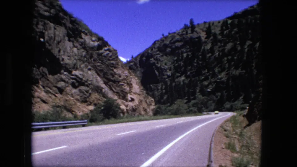 1976 home-movie frame from a moving car, showing a highway curve below rocky canyon walls and sparse trees.
