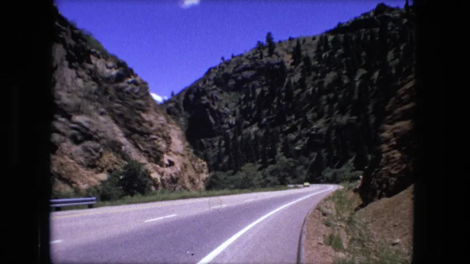 Cataloged 1976 8mm frame showing a two-lane highway bending through steep rocky terrain with trees along the slope.