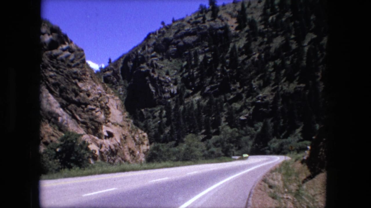 1976 8mm frame of a winding mountain highway seen from inside a vehicle, with rocky cliffs and trees along the road.