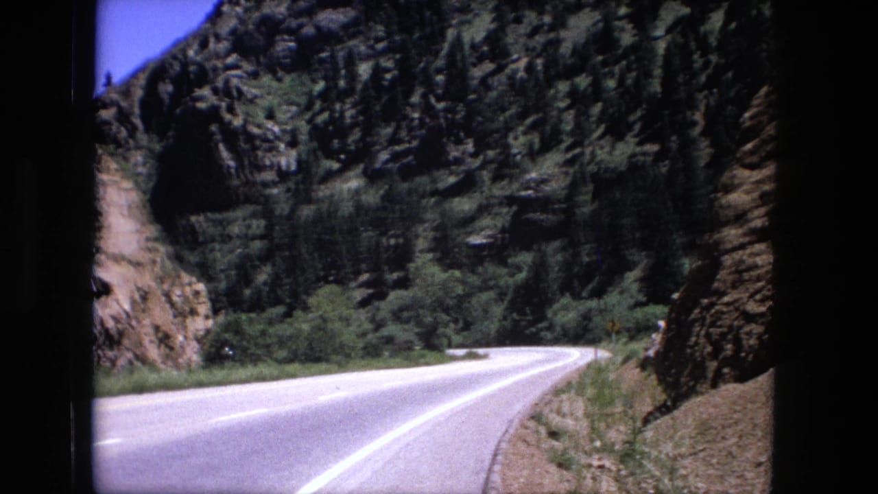 1976 8mm view of a paved highway bending through rocky hills, filmed through a dark vehicle window edge.