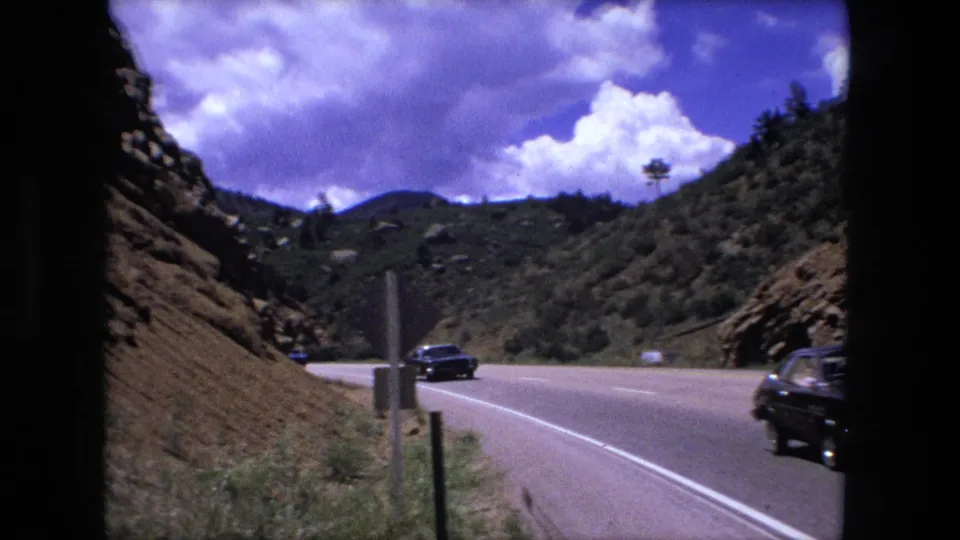 1976 8mm frame of cars traveling on a mountain highway, with roadside posts, hills, and clouds visible.