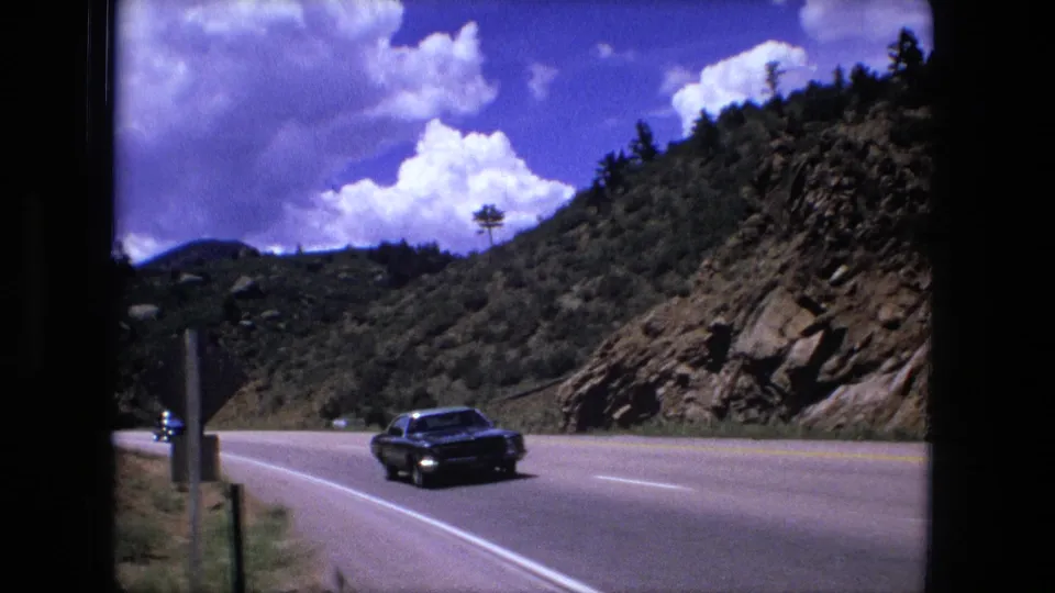 1976 8mm view of a dark sedan on a bright mountain road with rocky hillsides and clouds behind it.