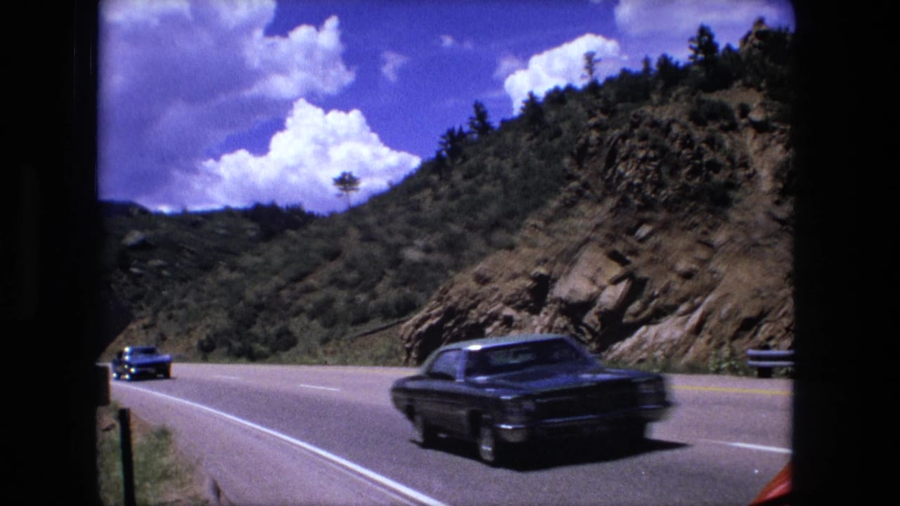 1976 8mm frame of cars traveling along a curving two-lane highway through a rocky mountain pass.