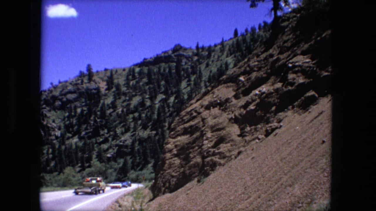 Several vehicles move through a highway curve below a rocky, tree-covered slope.