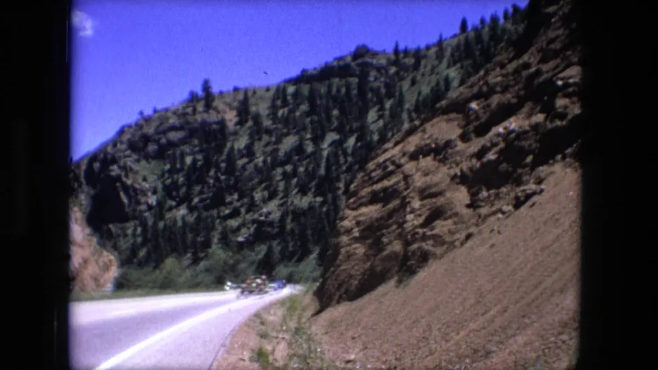 1976 8mm frame of a two-lane highway curving beside a rocky slope with cars visible in the distance.