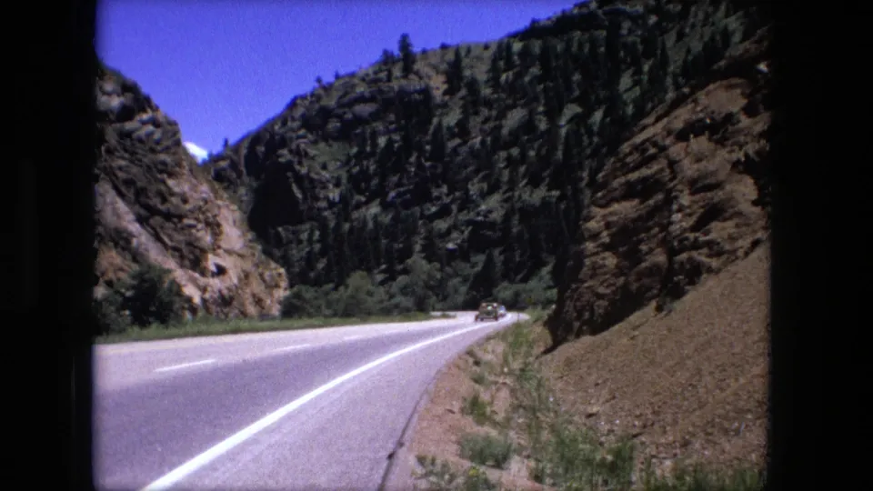 1976 8mm frame showing a car driving away along a mountain highway bordered by rock cuts and evergreens.