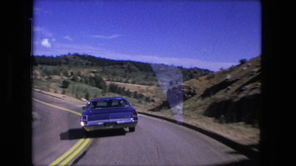 1976 8mm road-trip frame following a blue sedan on a two-lane mountain road with hills and open sky ahead.