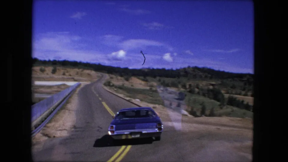 1976 8mm frame showing a blue sedan ahead on a two-lane road, with guardrail, low hills, and a bright sky in view.