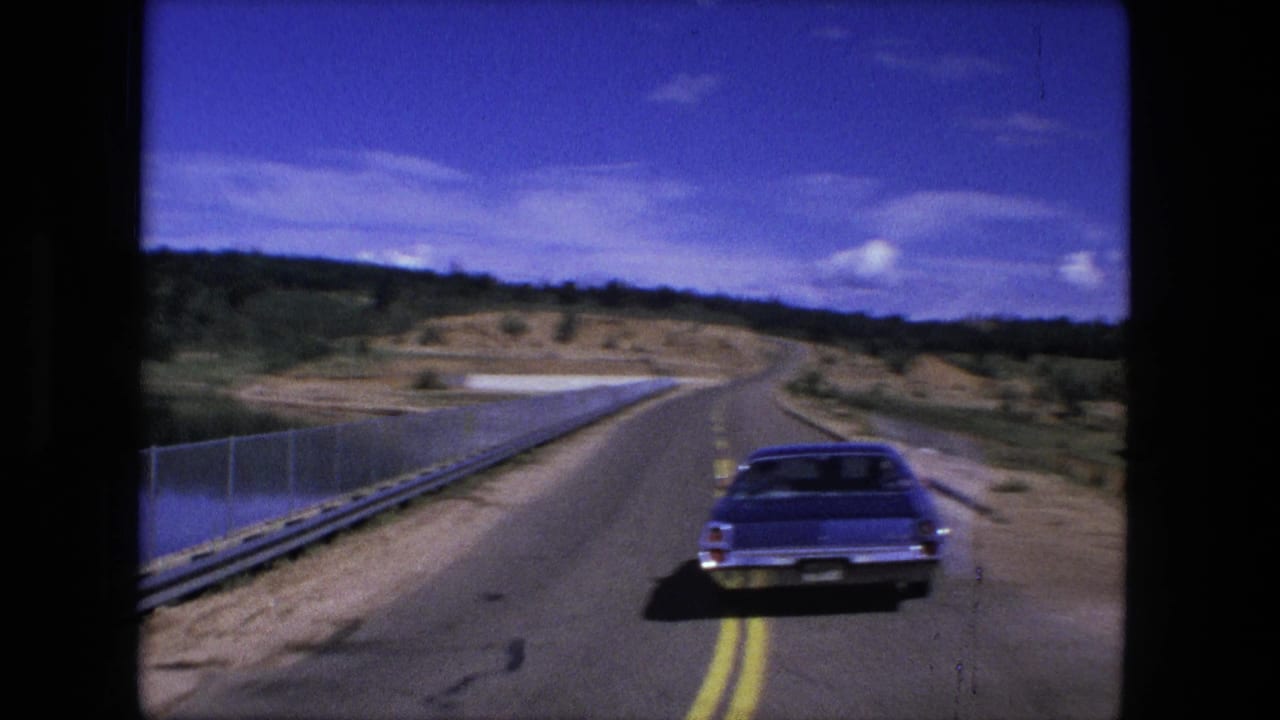 Blue classic car driving ahead on a paved rural road beside water and guardrails.