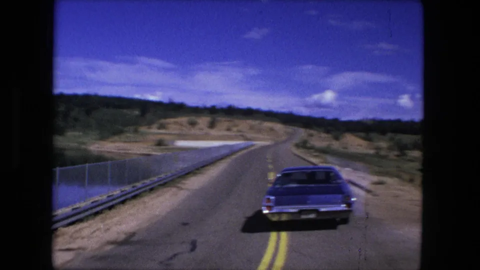 1976 8mm home-movie view of a blue classic car traveling on a rural road near water, with guardrails and open hills ahead.